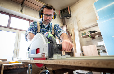 Woodworker wearing safety gear using a circular saw to cut a wood at woodworking studio