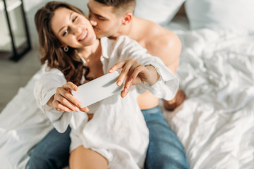 selective focus of cheerful girl taking selfie on smartphone while boyfriend kissing her