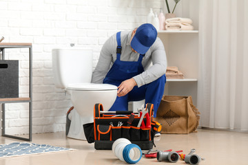 Handsome plumber repairing toilet bowl in bathroom - obrazy, fototapety, plakaty