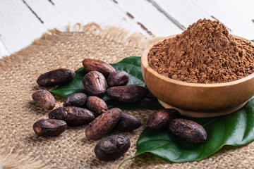 Cacao powder in wooden bowl and whole cacao beans with original fresh leaves on white rustic table.