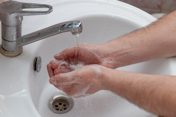 A man washes his hands in a white washbasin. An open tap, running water, soap suds on hand. Personal hygiene. Soap.
