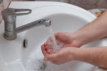 A man washes his hands in a white washbasin. An open tap, running water, soap suds on hand. Personal hygiene. Soap.