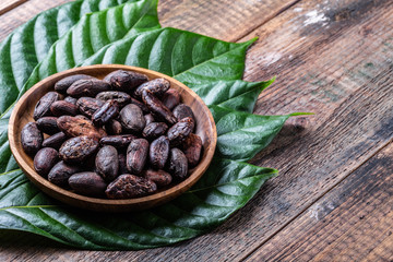Whole cacao beans in wooden bowl with original fresh leaves on rustic  table close-up.