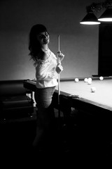 young woman with black hair in a white blouse stands near a poo,l table holds a cue in her hands, Brunette girl near the pool table,