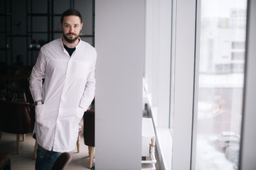Fototapeta premium Serious doctor with a beard stands by the window and looking at camera. Male doctor posing in modern meeting office room of the hospital. Concept of medical work.