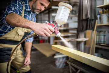 Carpenter with paint spray gun painting wooden plank in carpentry workshop