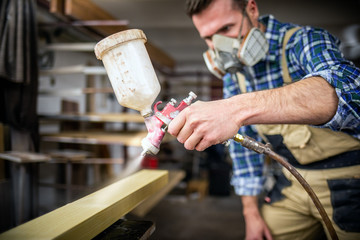 Carpenter with paint spray gun painting wooden plank in carpentry workshop