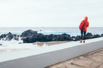 Tourist woman with backpack enjoying the power of ocean looking at waves surf at Porto Moniz natural pools, Madeira island, Portugal.