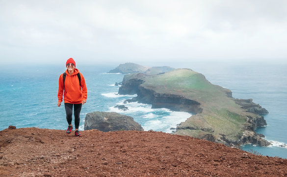 Young female backpacker dressed orange sporty hoodie and red cap hiking and enjoying Atlantic ocean view on Ponta de Sao Lourenço peninsula -the easternmost point of Madeira island, Portugal