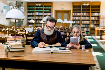 Cute little girl uses the application on the tablet, and an elderly man reads a book in the library. Old and new technologies, paper book and ebook, grandfather and granddaughter