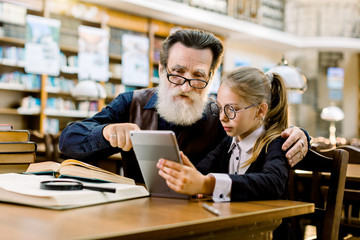 Senior man grandfather and little cute girl granddaughter looking at a tablet, while sitting at the table in old stylish library