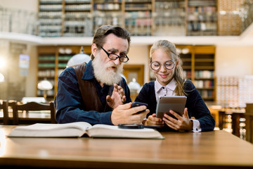 Grandfather and granddaughter, teacher and student, sitting together at table using digital tablet, smartphone and books. Study, family reading in library. Education concept