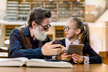 Handsome senior man and little cute girl sitting together in vintage library, compare books, smartphone and new reading digital book device. Grandfather and granddaughter in library