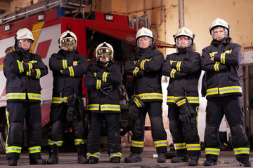 Six professional firefighters posing together. Firefighters wearing uniforms and protective helmets. Firetruck in the background.