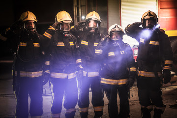 Group of professional firefighters posing. Firemen wearing uniforms, protective helmets and oxygen masks. Smoke and firetrucks in the background.