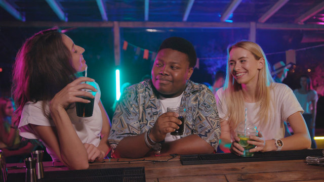 Two attractive girls meeting up approaching to handsome african boy. Happy young man ordering cocktail drinks for his women at modern night party.