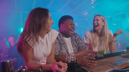 A couple of charming happy girls and positive african american guy laughing telling jokes chilling together at bar counter. Party and summertime.