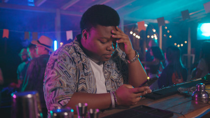 Tired and lonely afro-american guy checking social media on mobile phone covering face suffering of boredom staying at bar night modern party. Social life concept.