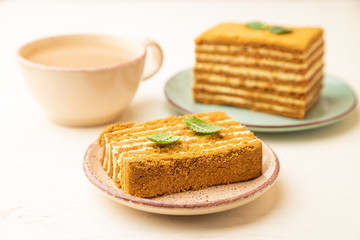 Homemade honey cake with milk cream and mint with cup of coffee on a white wooden background. Side view, selective focus.