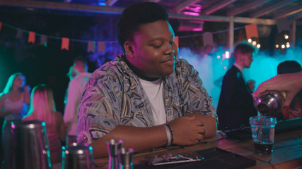 Afro-american young man in stylish look sitting on bar enjoying music with headshake using his modern smartphone on night party.