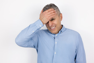 adult handsome man with grey hair wearing blue shirt touching forehead with hand feeling sorry standing on isolated white background