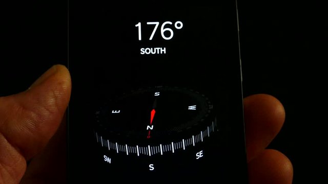 Fingers holding a mobile phone with a compass app displayed, showing the north, south, east, west direction pointer and the geographic decimal degrees as the compass moves.