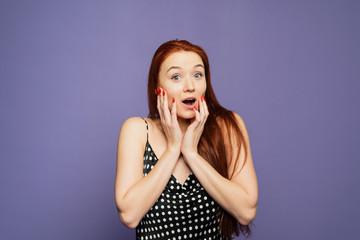 Surprised happy young female stands on purple background, glad to see big discounts on clothes, expresses shock, keeps hands near head, jaw dropped, going to spend much money today