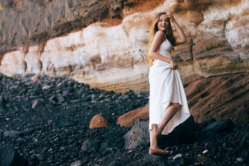 Gorgeous girl in a long white dress on the ocean at the beach. The long-haired girl is inherited by nature, mountains, stones, the beach. Girl on vacation, light dress, summer, date. Bride