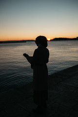 Fototapeta premium Silhouette of young European Muslim woman with hijab walking by the sea during sunset/sunrise time. Winter time.