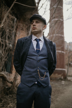 A man posing in the image of an English retro gangster dressed in Peaky blinders style near abandoned building.