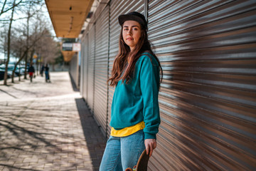 Young beautiful hipster woman posing with her hand on a skateboard. In the background, a street with closed shops. Cloe up. Concept of sports lifestyle and street culture