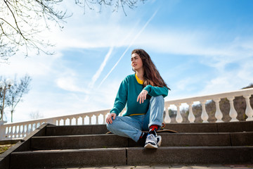 Obraz premium Caucasian teenage girl sitting on a skateboard and looking into the distance.In the background, the blue sky and the Boulevard.Bottom view.Concept of sports lifestyle and street culture