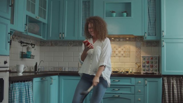 Smiling curly 20s woman in jeans dancing happily with scoop as microphone and mobile phone in hands on home blue kitchen. 