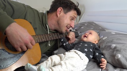 An attractive father playing guitar to his adorable baby boy on a bed