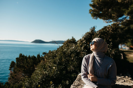 portrait of young European Muslim women with hijab sitting on the stone beach with sea and port in the background. She is happy and enjoying sun.