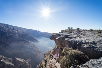 Man peeking over the cliff with the sun in the background at the Grand Canyon of Jebel Shams, Oman