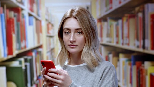 Caucasian pretty girl young student using a smartphone social network app messaging while studying at bookshelves in the library. Modern education concept.