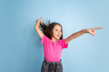 Pointing on in jump. Caucasian little girl's portrait on blue studio background. Beautiful female model in pink shirt. Concept of human emotions, facial expression, sales, ad, youth, childhood.