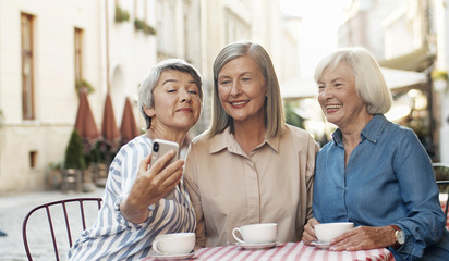 Three Caucasian pretty senior women friends sitting with coffee at table in cafe terrace and smiling to smartphone camera while taking selfie photos. Old ladies posing to phone. Pensioners resting.