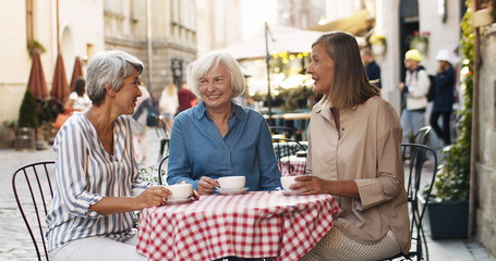 Three Caucasian senior women friendly chatting and drinking coffee at table in cafe terrace in...