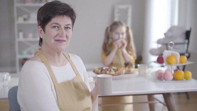 Positive brunette Caucasian woman drinking tea and smiling at camera as blurred little girl eating pancakes at the background. Happy grandmother spending Shrove Tuesday with granddaughter.