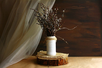 Willow branches in a white vase on a wooden stand cut, chiffon beige golden tulle on a dark brown background. The interior is in a rustic pastoral eco style.