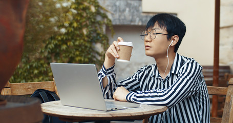 Asian guy in glasses and headphones sitting at table in cafe terrace and drinking coffee. Young man having rest while working on laptop computer. Chinese male student sipping drink and resting.