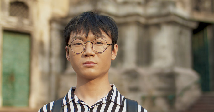 Close Up Of Asian Young Good Looking Straight To Camera Man Outdoor At Antique Old Building. Portrait Of Male Student With Serious Straight Outdoors. Chinese Guy Traveller In Glasses.