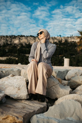 portrait of young European Muslim women with hijab sitting on the stone beach with sea in the background. She is happy and relaxed.