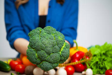 A woman at a table holding a broccoli on a background of fruit and vegetables