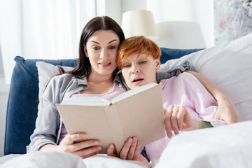 Excited woman reading book near girlfriend on bed