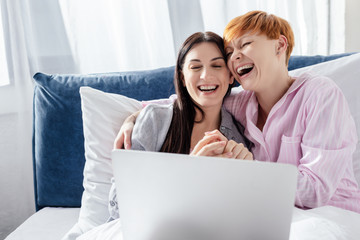 Same sex couple laughing while using laptop on bed