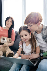 Selective focus of woman hugging daughter while using laptop near parent with teddy bear on couch