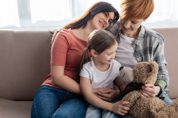 Smiling same sex parents sitting near daughter with teddy bear on couch